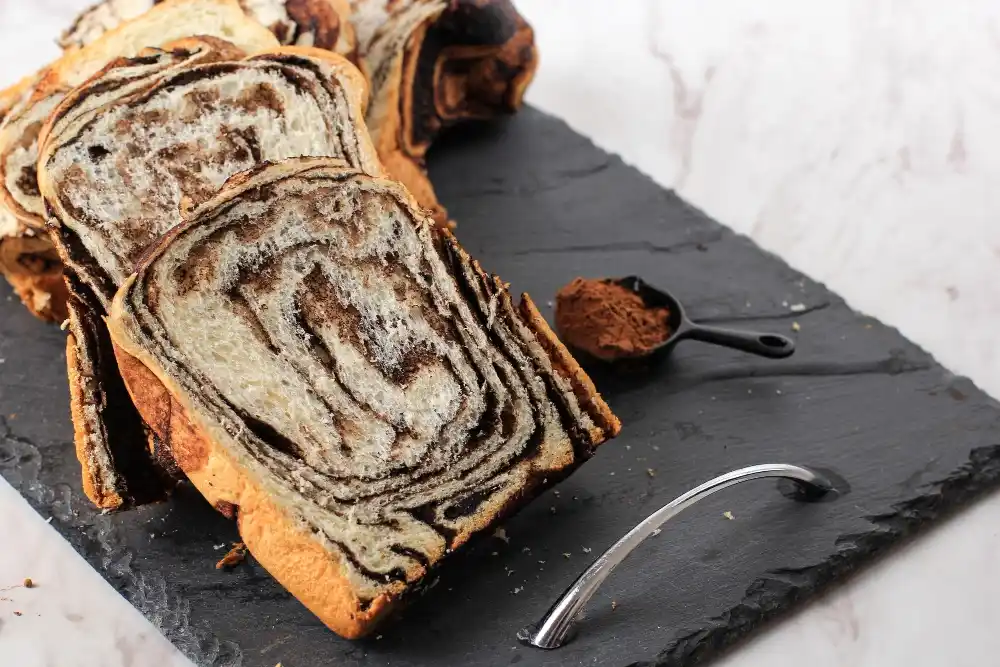 Banana Bread Cinnamon Rolls: A Step-by-Step Guide 4 Loaf of marbled banana bread with cinnamon swirls, sliced and arranged on a slate serving board, accompanied by a spoonful of ground cinnamon and a decorative silver utensil.