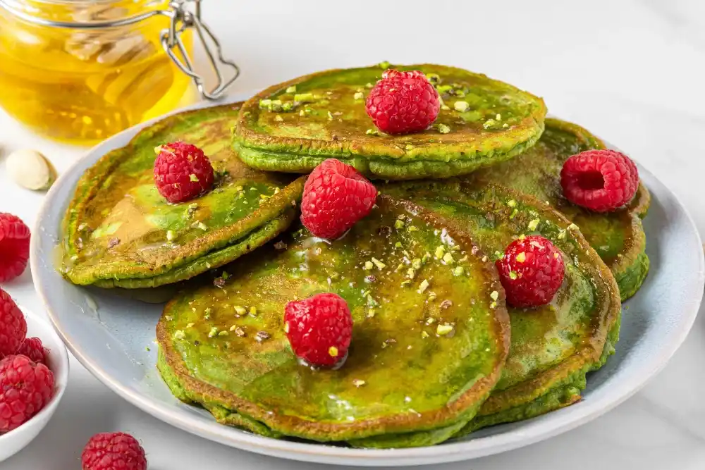 Mini Protein Pancakes Recipe - Healthy and Tasty Breakfast Option 3 A stack of vibrant green pancakes garnished with fresh raspberries and crushed pistachios, drizzled with honey, served on a light blue plate with a jar of honey and additional raspberries in the background.