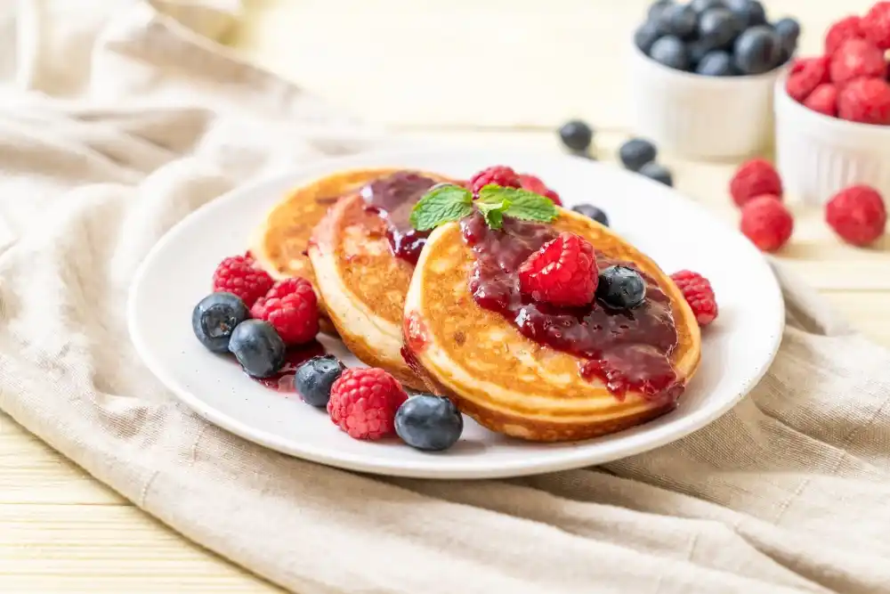 fluffy pancakes topped with berry sauce, fresh raspberries, blueberries, and a sprig of mint, served on a white plate with additional berries in the background on a light wooden table.